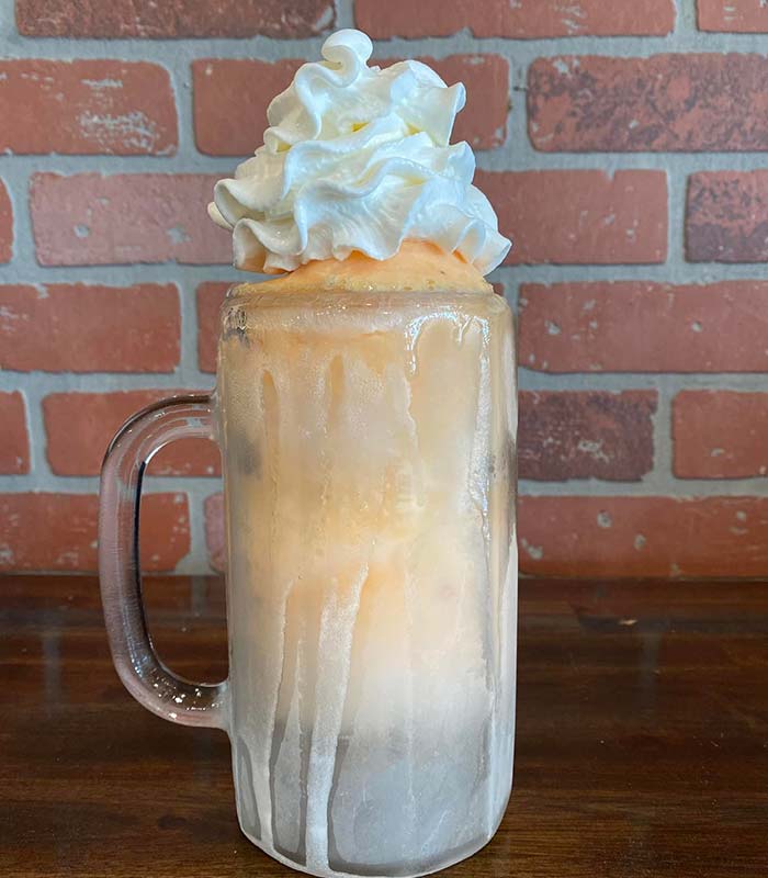 A frosty glass mug filled with a light orange ice cream float, topped with a swirl of whipped cream, sits on a wooden surface at Gatesville Bar, adding charm to the cozy brick wall backdrop.