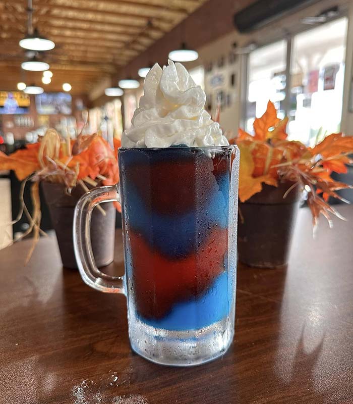 A frosty glass mug filled with a layered blue and red drink, topped with whipped cream, sits on a wooden table at Studebakers Pizza & Tap Wall Gatesville TX. Two potted plants with orange autumn leaves add charm to the cozy café scene.