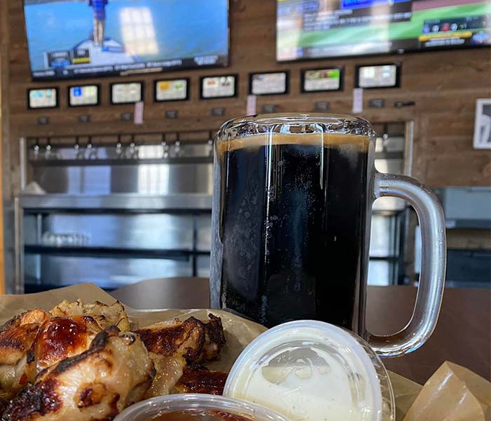 A glass of dark beer sits on a table at Studebakers Pizza & Tap Wall Gatesville TX, next to saucy chicken wings and ranch. In the background, beer taps and TV screens elevate this classic Gatesville Bar experience.