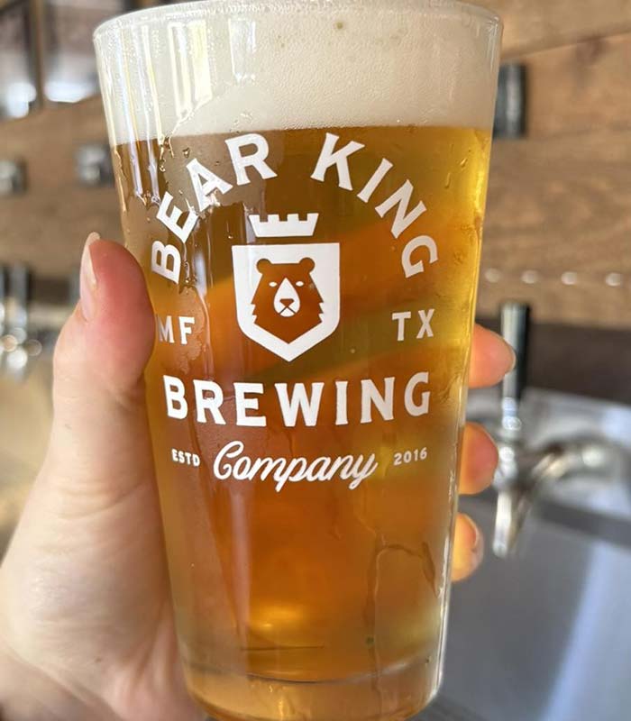 A hand holds a pint glass of light beer labeled Bear King Brewing Company, with a bear logo and MF TX Est. 2016. Beer taps blur in the background at Studebakers Pizza & Tap Wall Gatesville TX, a popular Gatesville restaurant and bar.