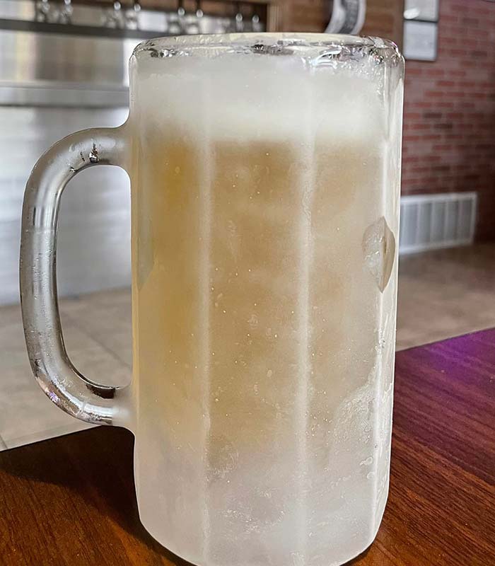 A frosty glass beer mug filled with light beer, condensation and ice coating the outside, sits on a wooden table at Studebakers Pizza & Tap Wall Gatesville TX, with a metal counter and brick wall in the background.