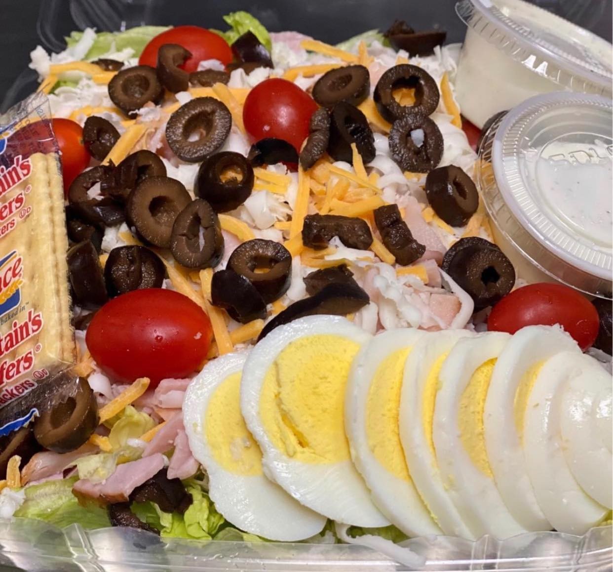 Enjoy a fresh salad from Gatesville Restaurant, featuring sliced hard-boiled egg, cherry tomatoes, ham, shredded cheese, black olives, crisp lettuce, crackers, and two dressings—all in a convenient clear takeout container.