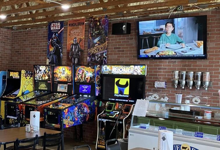A retro arcade with pinball machines, movie posters, and a TV showing a boy eating pizza brings nostalgia to Studebakers Pizza & Tap Wall Gatesville TX. An ice cream freezer sits by a brick wall; tables and chairs are partially visible in the foreground.