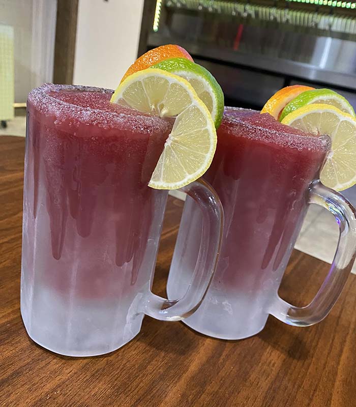 Two frosty glass mugs filled with a red frozen drink, garnished with lime and orange slices, sit on a wooden table at Studebakers Pizza & Tap Wall Gatesville TX—your go-to Gatesville Bar for refreshing treats.