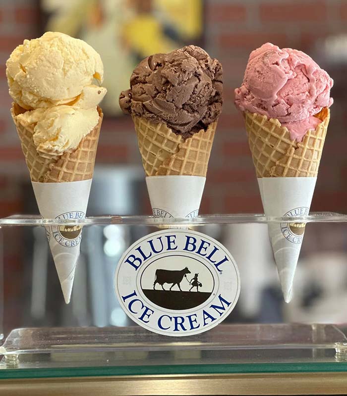 Three waffle cones with vanilla, chocolate, and strawberry ice cream stand upright in a holder featuring the Blue Bell Ice Cream logo—a nod to the sweet treats you’ll find at Gatesville Pizza, a favorite Gatesville restaurant.