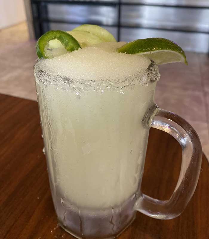 A frosty glass mug filled with a frozen margarita, topped with two lime wedges and a jalapeño slice, sits on a wooden table at Studebakers Pizza & Tap Wall Gatesville TX. The rim of the glass is coated with salt.