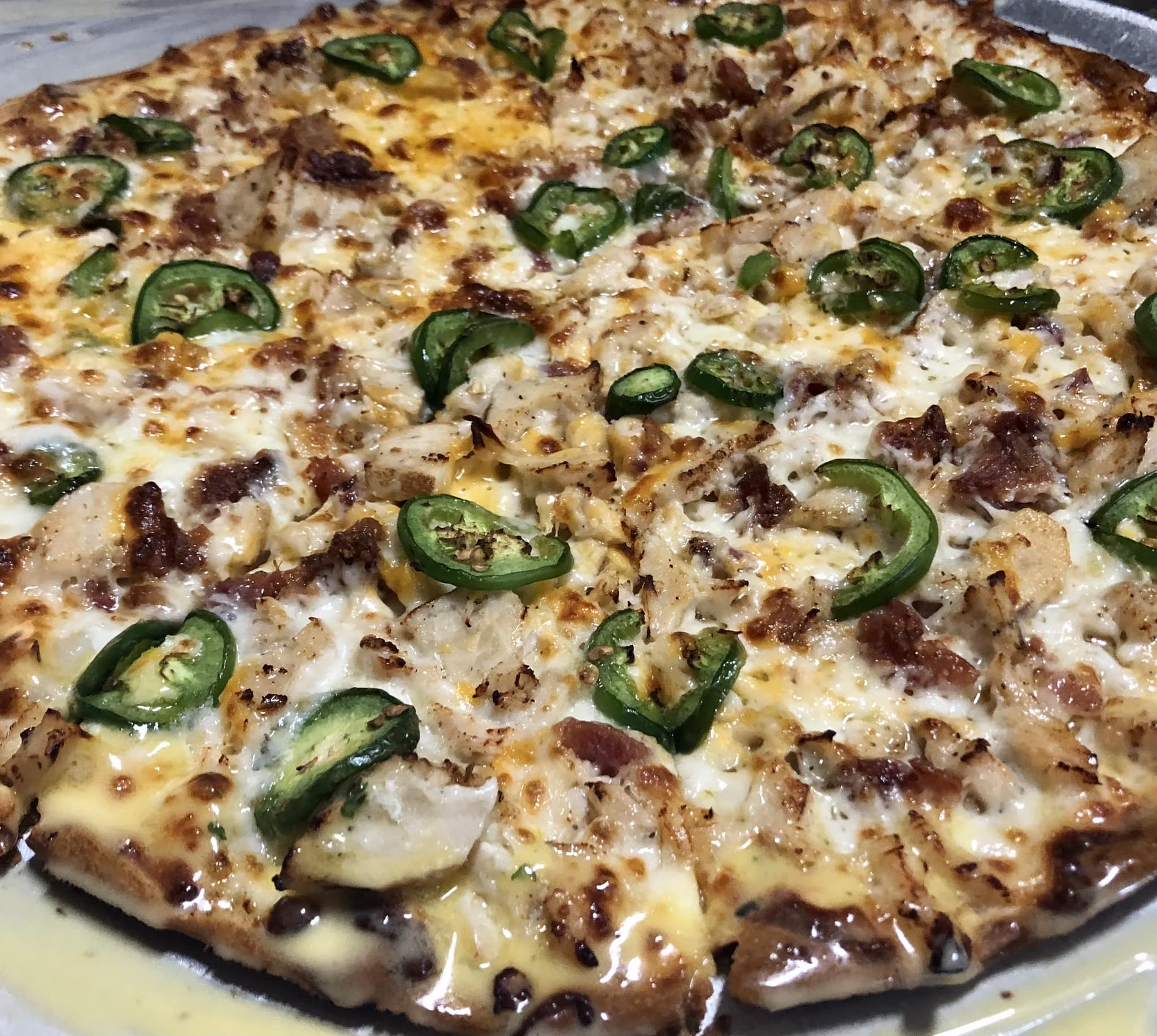 A close-up of a baked Gatesville Pizza topped with melted cheese, sliced jalapeños, chunks of chicken, and bits of bacon on a thin crust at Studebakers Pizza & Tap Wall Gatesville TX.