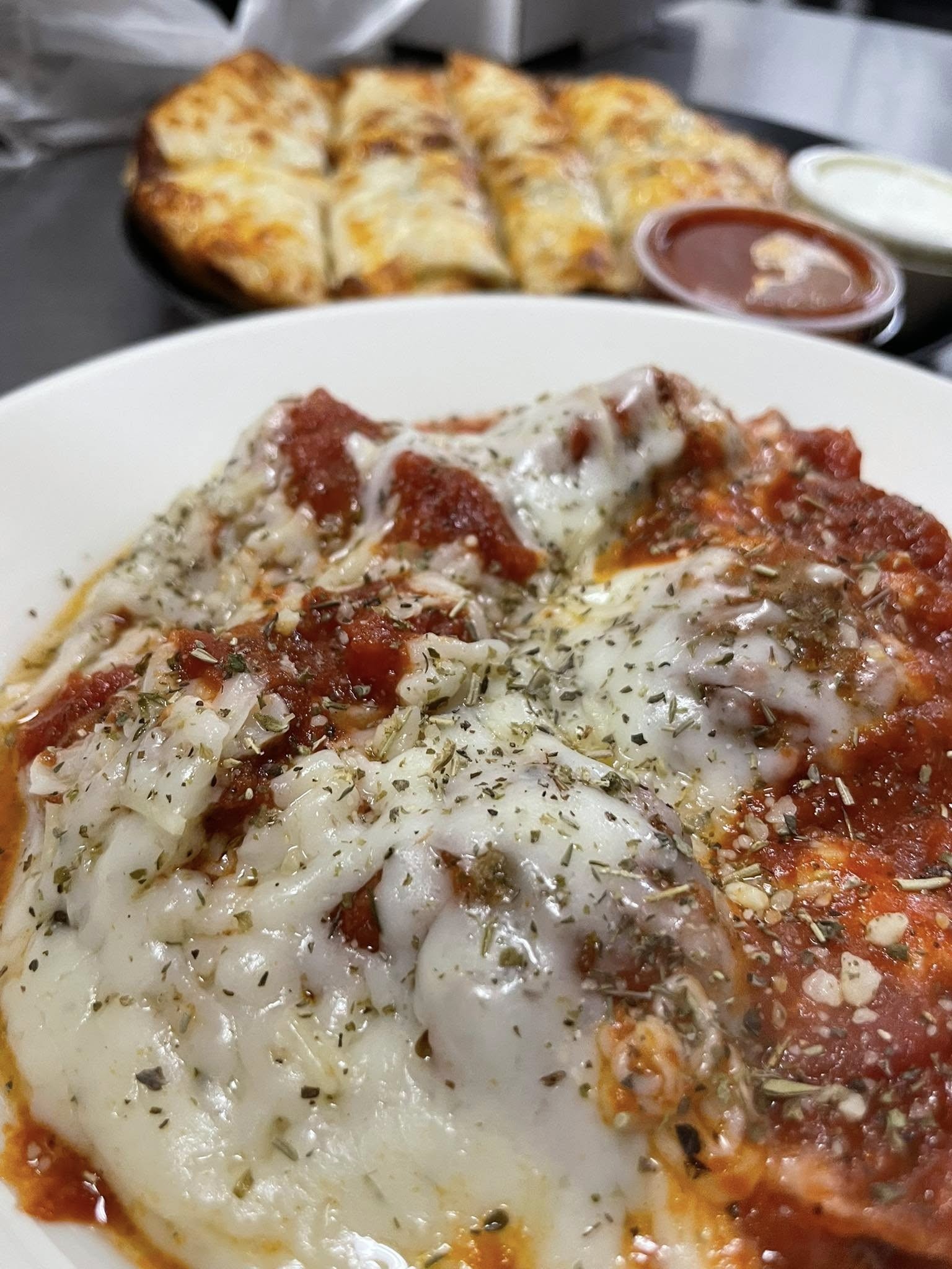 A close-up of cheese-covered ravioli topped with tomato sauce and herbs in a white bowl, with a Gatesville Pizza and dipping sauces in the background at Studebakers Pizza & Tap Wall Gatesville TX.
