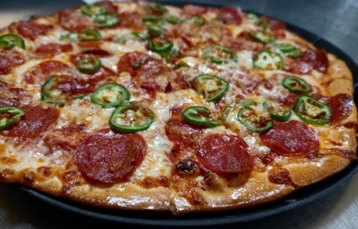 A freshly baked pizza topped with melted cheese, pepperoni, and jalapeño peppers on a golden-brown crust, served on a round black tray at Studebakers Pizza & Tap Wall Gatesville TX—a favorite spot for pizza lovers in Gatesville.