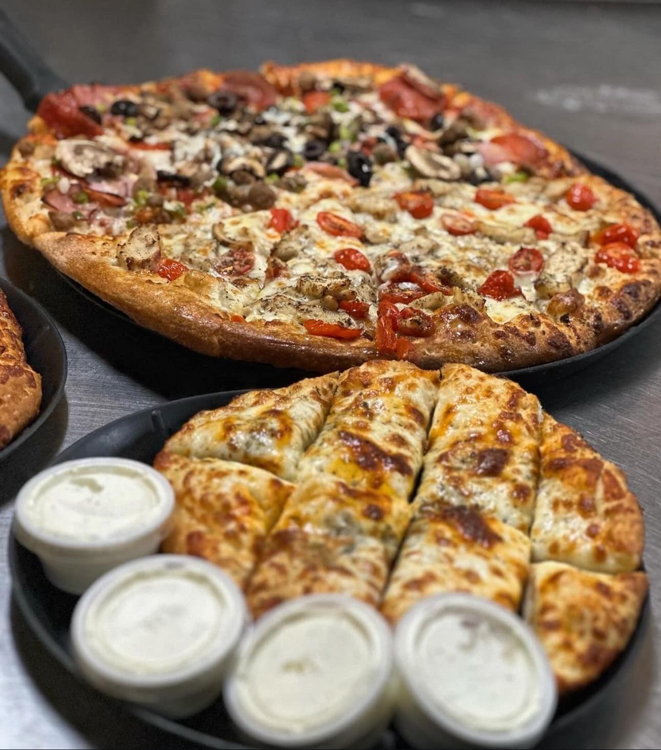 A large Gatesville Pizza topped with vegetables and meat sits behind cheesy breadsticks, served with three cups of dipping sauce on a metal surface at Studebakers Pizza & Tap Wall Gatesville TX.