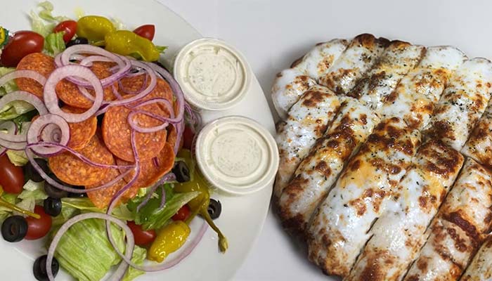 A plate with a fresh salad topped with pepperoni, red onions, olives, and tomatoes plus two cups of ranch sits next to cheesy breadsticks—just one tasty option at Gatesville Pizza, your favorite Gatesville Restaurant.