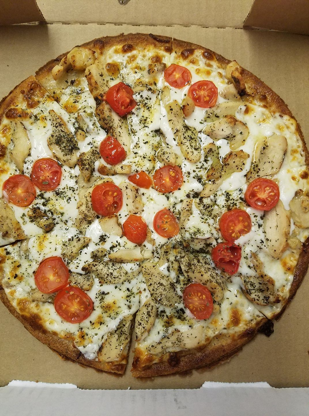 A Gatesville Pizza in a cardboard box topped with grilled chicken strips, melted cheese, halved cherry tomatoes, and sprinkled herbs. The pizza is sliced and features a golden-brown crust—perfect for any Gatesville Restaurant fan.
