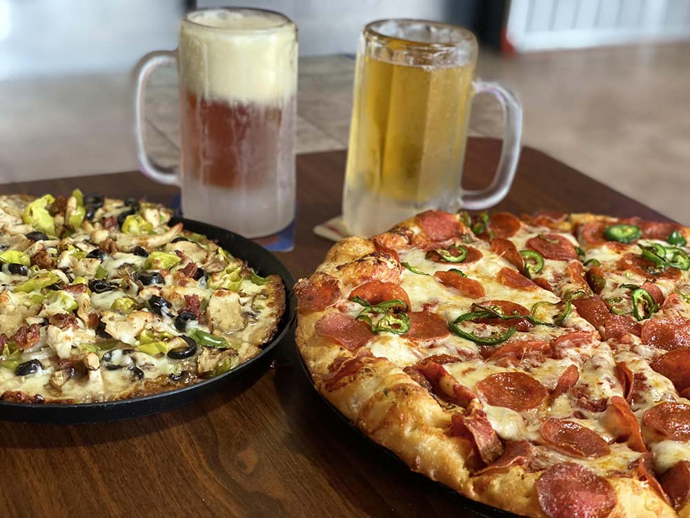 Two mugs of beer sit behind two pizzas at Studebakers Pizza & Tap Wall Gatesville TX; one pizza is topped with pepperoni and jalapeños, while the other features veggies like onions, black olives, mushrooms, and pepperoncini peppers.