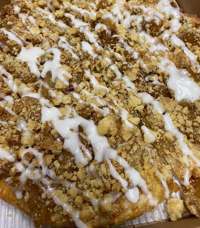 A close-up of a dessert topped with apple chunks, crumbly streusel, and drizzled with white icing, served fresh at Studebakers Pizza & Tap Wall Gatesville TX, giving it a sweet and textured appearance.