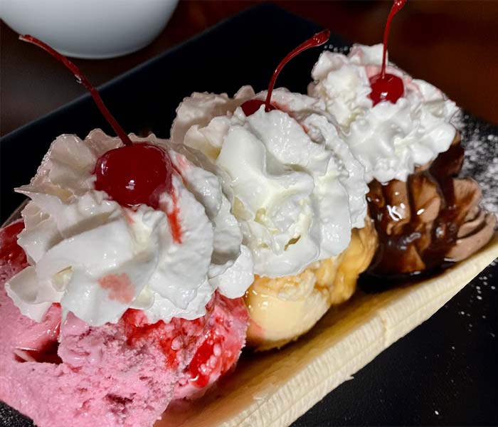 A banana split with scoops of strawberry, vanilla, and chocolate ice cream, each topped with whipped cream and a cherry, served on a black plate at Gatesville Restaurant.