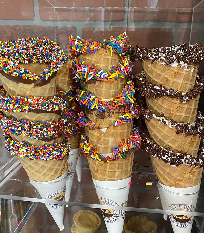 Waffle ice cream cones dipped in chocolate and decorated with colorful rainbow and chocolate sprinkles are displayed upright in a glass case at Gatesville Pizza, set against a charming brick wall background.