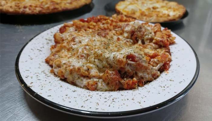 A plate of baked pasta topped with melted cheese and herbs sits on a metal surface, with two pizzas blurred in the background at Studebakers Pizza & Tap Wall Gatesville TX.