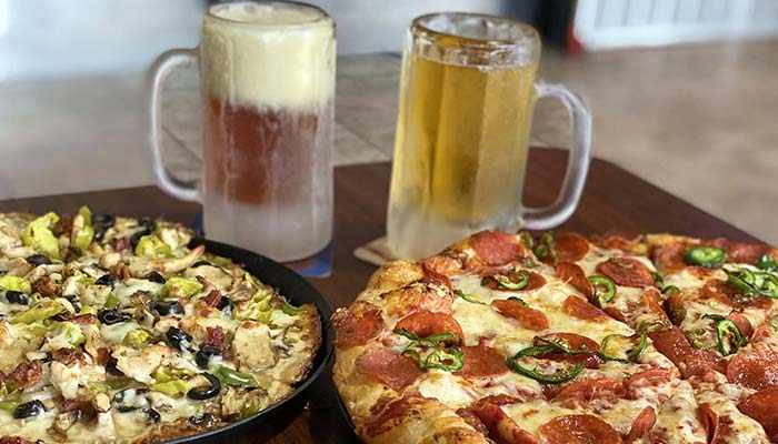Two frosty mugs of beer sit behind two large pizzas at Studebakers Pizza & Tap Wall Gatesville TX; one pizza is topped with pepperoni and jalapeños, the other with chicken, olives, green peppers, and mushrooms.