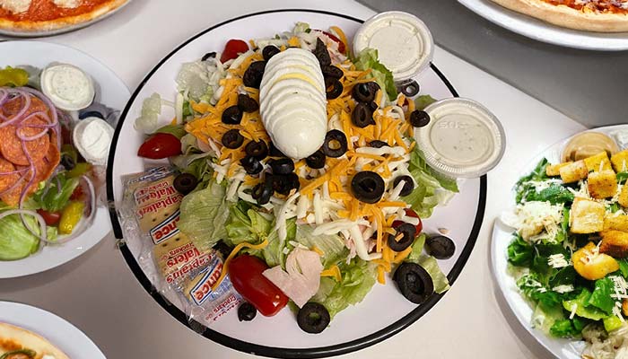 A chef salad at Gatesville Pizza, topped with sliced hard-boiled egg, shredded cheese, black olives, cherry tomatoes, deli meat, and dressing, served with crackers. Other salads and pizzas are partially visible around it.