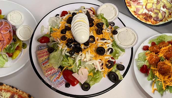 A large salad with sliced hard-boiled egg, shredded cheese, black olives, lettuce, tomatoes, dressing containers, and crackers on a white plate, surrounded by other plates of salad and Pizza and Wings Gatesville TX favorites.